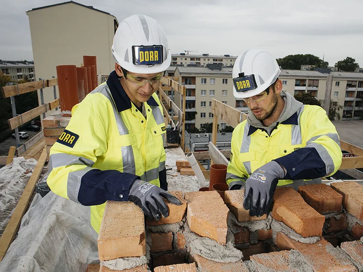 Two young PORR employees are on duty at a building construction project during their bricklayer apprenticeship. For their safety they are wearing helmets, the blue and yellow workwear and safety gloves. As a team they are working highly focused on the completion of a house. They are just putting bricks onto fresh mortar.