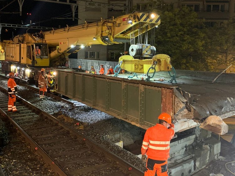 Excavation of the old bridge element Photo: The old bridge element is lifted with a crane. The employees in orange uniforms stand on the tracks and watch the procedure.