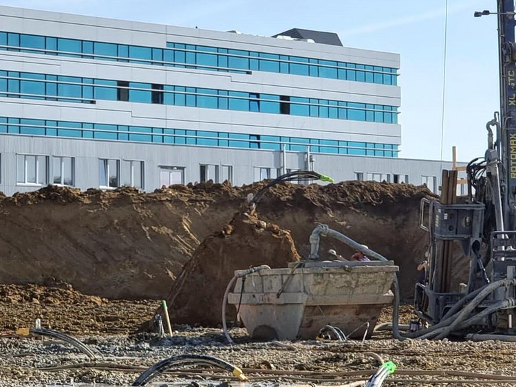 On the trail of heat. In the foreground, a construction site with a geothermal drilling rig and a skip, with pipes protruding from the ground. In the background is the existing FH Campus Wien building.