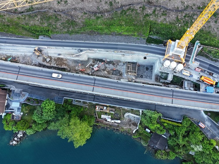 Excavations valley-side structures Photo: Aerial view of the construction site with large crane. The road is used by cars and is located between the lake and the steep rock face.