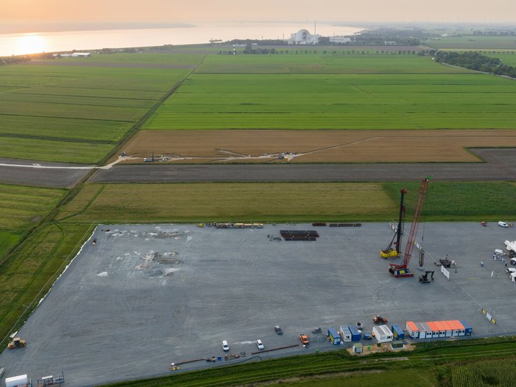 SuedLink Elbe crossing ElbX, Wewelsfleth / Wischhafen Aerial view of many green fields next to a large river. A large grey construction site can be seen in the middle of the fields, with the first construction containers, a pile driver and a diaphragm wall grab.